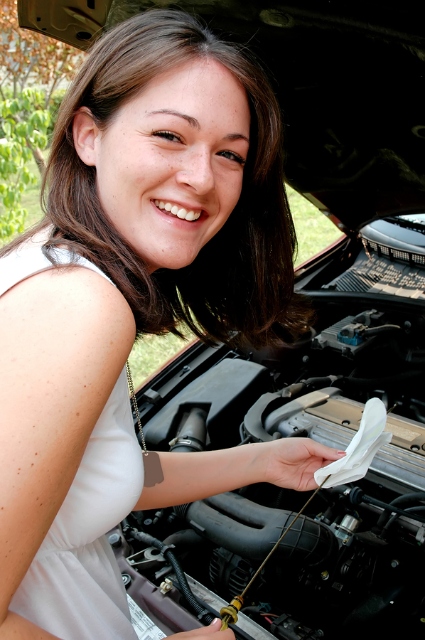 A woman grins as she wipes off a dipstick before an open car hood.